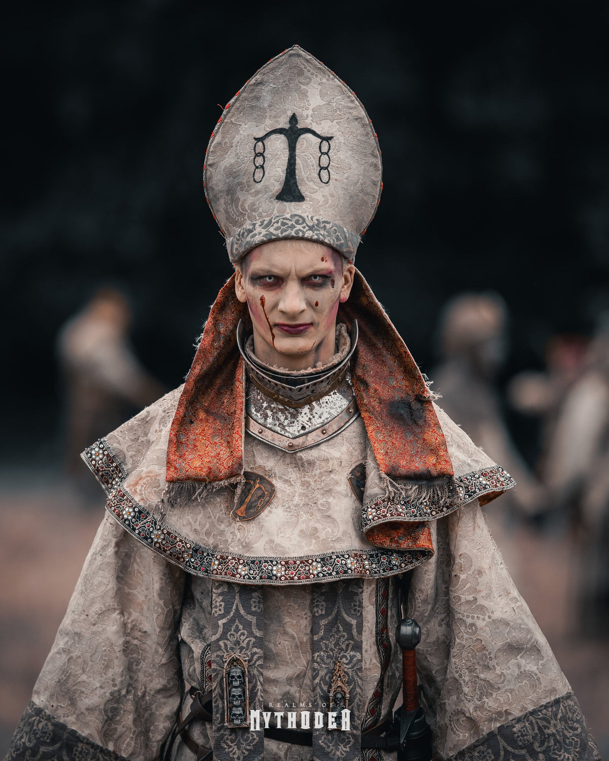 A person in elaborate ancient Egyptian or Egyptian-inspired ceremonial costume, including a tall headdress with a black scale symbol, a patterned robe, and an orange-brown neck drape, photographed against a dark blurred outdoor background with other blurred figures. Undead Flesh Mythodea LARP Burgschneider