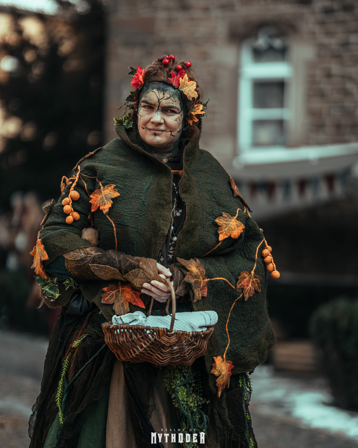 A person wearing a detailed autumn-themed costume stands outdoors holding a wicker basket. The costume includes a dark green hooded cloak decorated with orange and brown leaves, small orange berries, and a floral crown with red and orange blossoms. Mythodea LARP. 