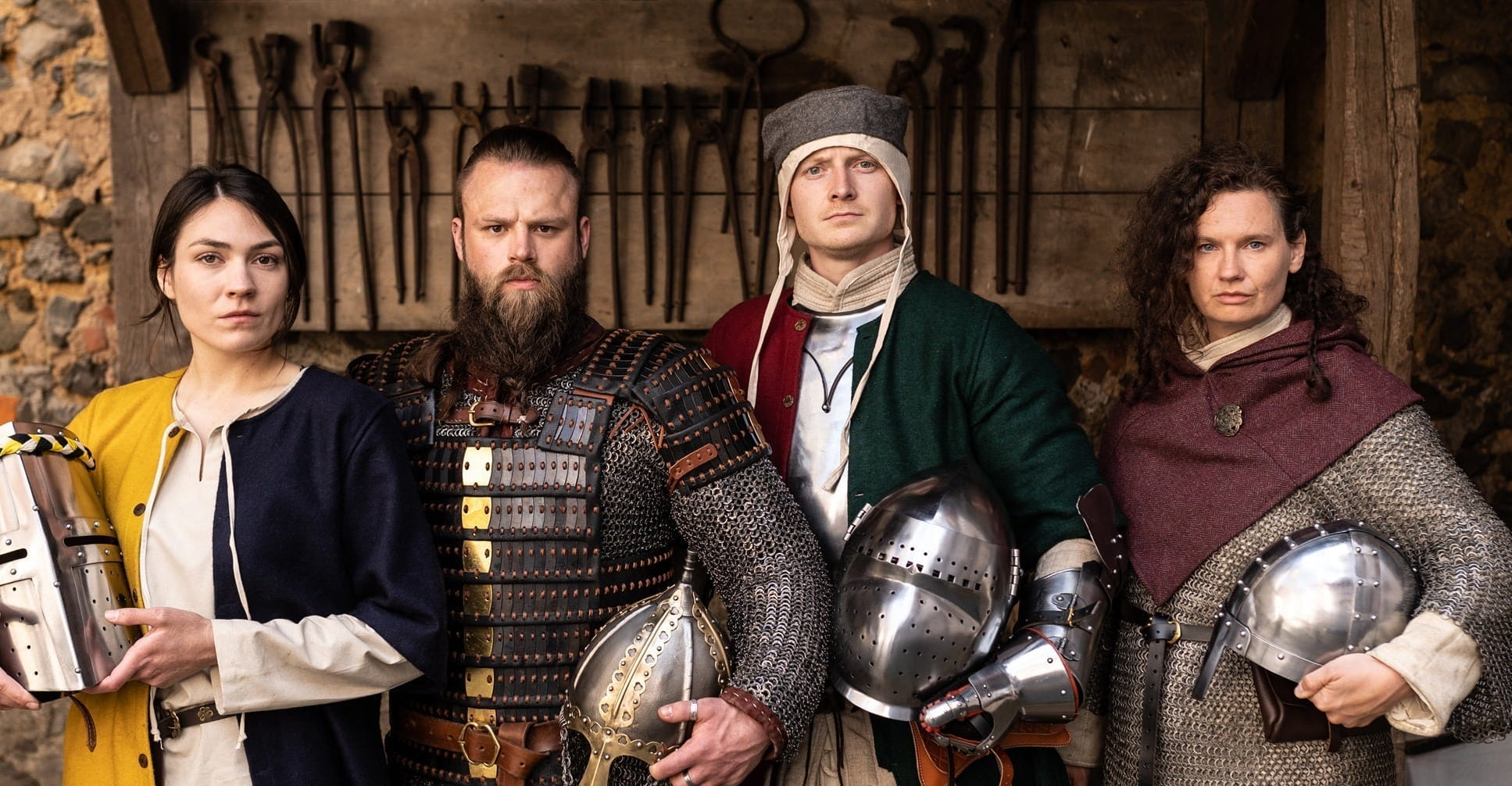 Four people in historical medieval clothing wearing armor and holding medieval helmets