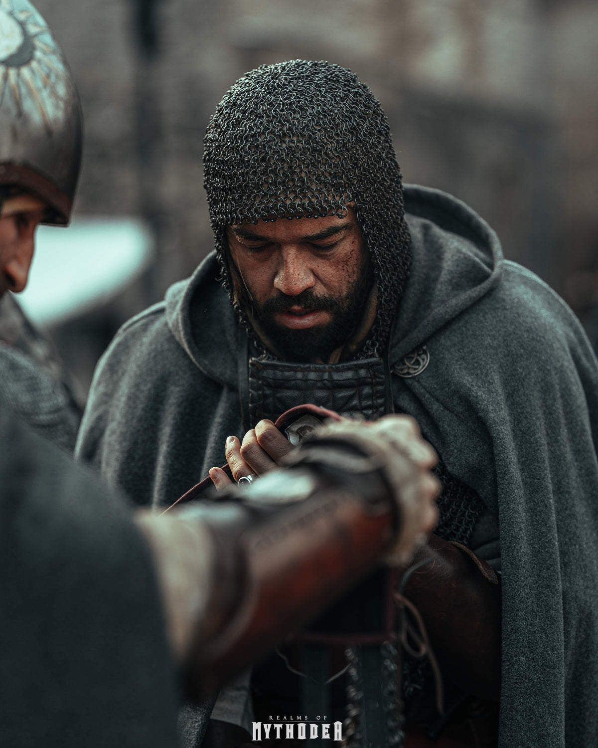 A man in medieval-style chainmail armor and a gray hooded cloak looks down while another person in a helmet is partially visible at the left edge. Mythodea Larp Burgschneider