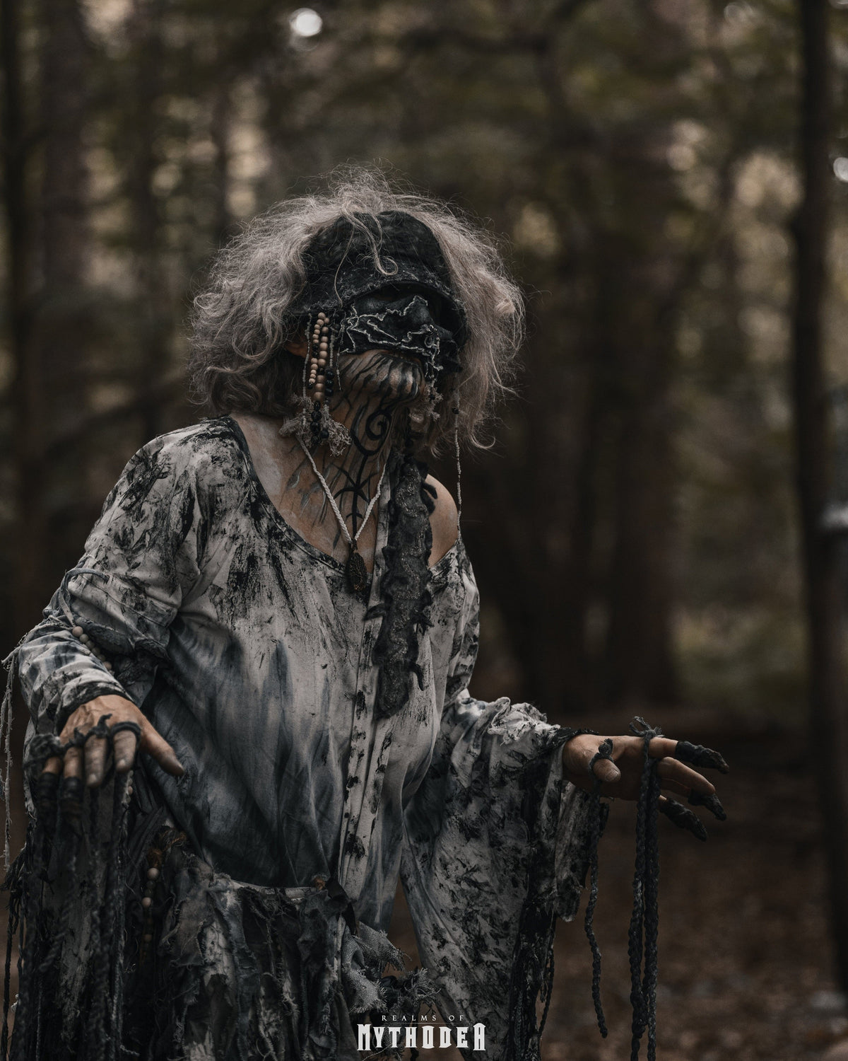 A person in a dark, distressed costume stands in a forest, wearing a tattered black and gray dress, a black head covering with beaded details, and holding dark string. The background is a blurred wooded setting. Mythodea Larp Burgschneider