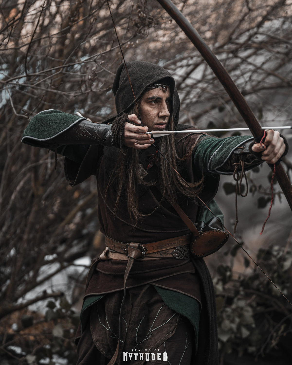 A person in medieval-style dark green and brown robes draws a bow in a wooded outdoor setting. Mythodea Larp Burgschneider