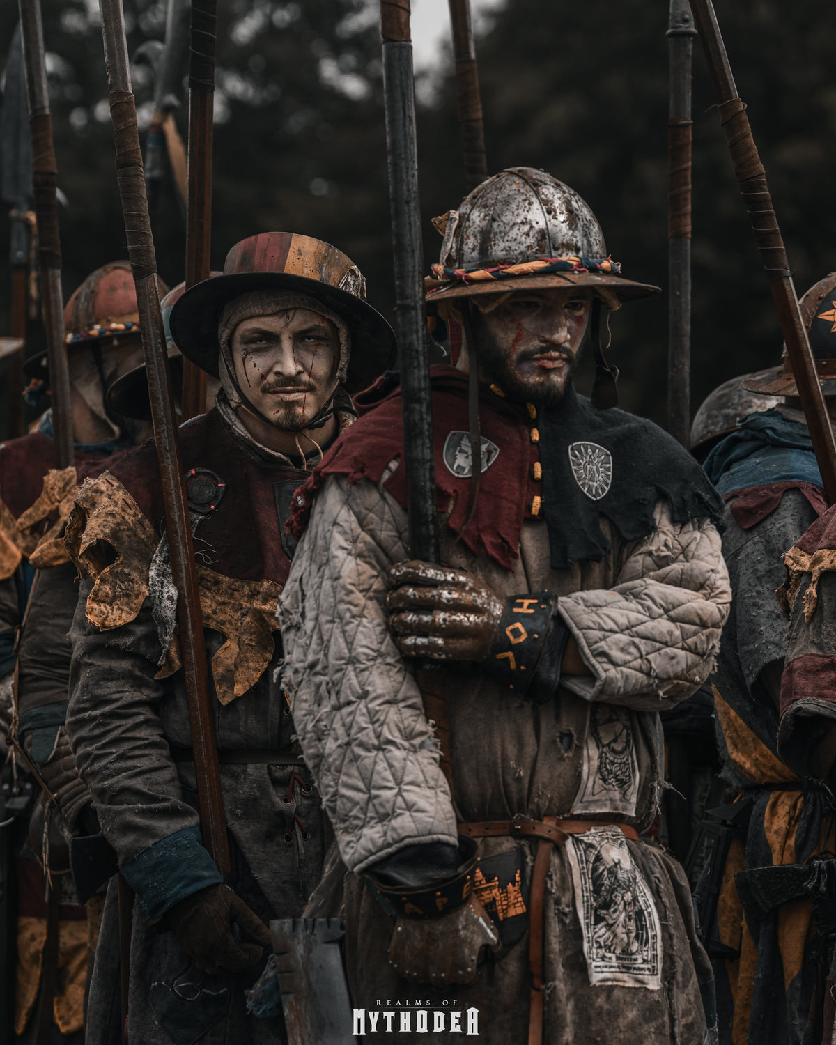 Mythodea Larp Burgschneider. A group of fantasy warriors in detailed medieval-style armor and helmets, holding long spears in a dark, atmospheric outdoor setting. The central figure wears a weathered metal helmet, a quilted gray tunic, and a dark red cape.