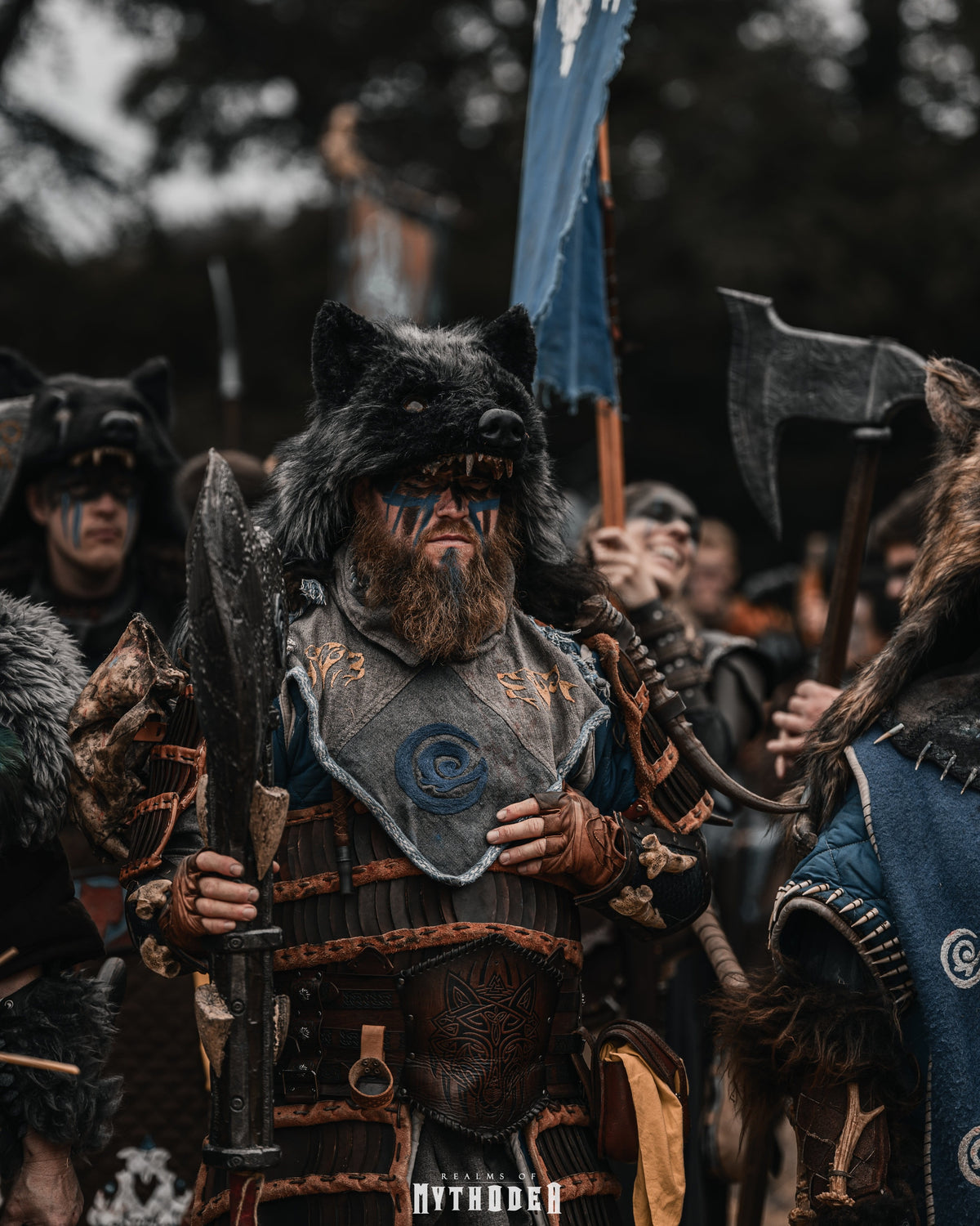 Mythodea Larp Burgschneider. A person in a Viking-style costume stands in a crowd, wearing a wolf mask, dark fur, painted face, and detailed armor with a blue shield-like chest piece. Axes and other participants are visible in the background and around the subject.