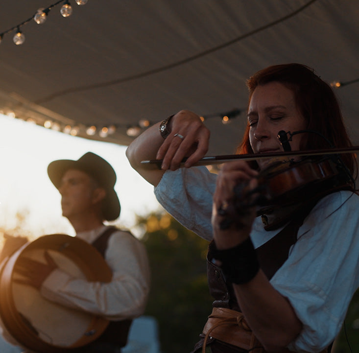 Person playing violin with another person playing banjo under a tented outdoor setting.
